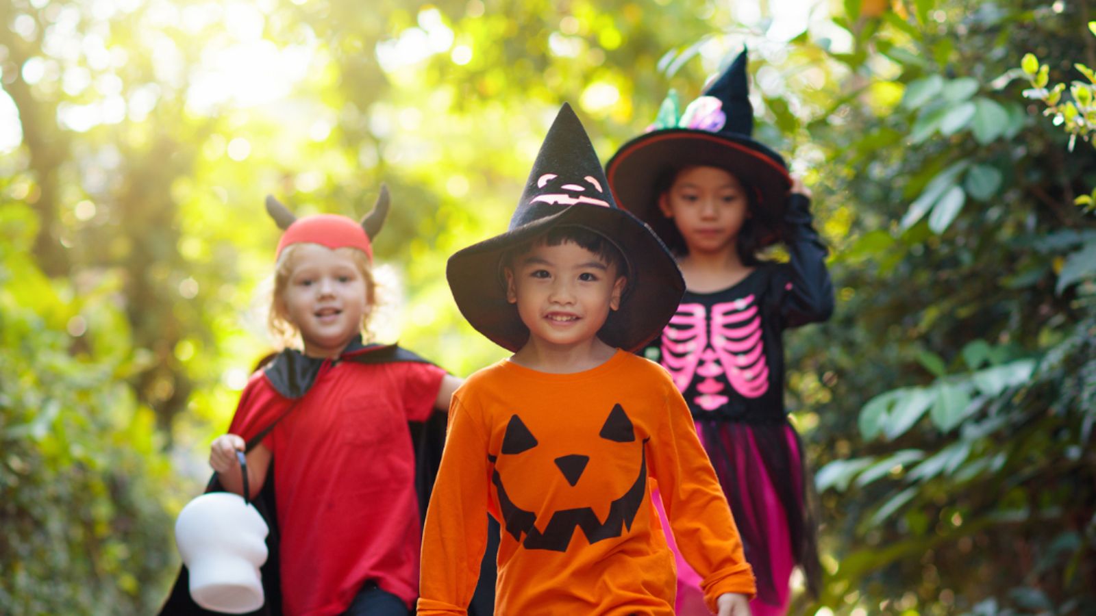 Three children walking towards the camera in halloween costumes, one is dressed as a devil - carrying a trick or treat basket, one has a pumpkin shirt on with a witches hat, and one is dressed as a witch. 